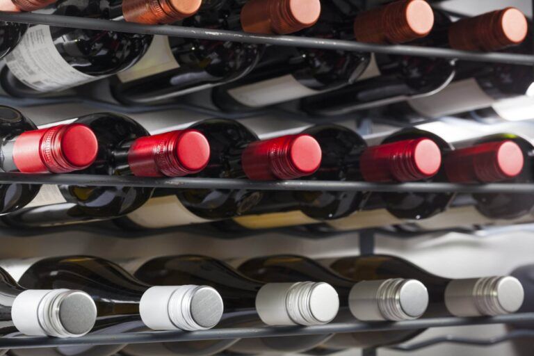 Close-up of numerous wine bottles stored horizontally in tilted wine racks in a cellar, featuring a variety of caps including red, white, and metallic colors.