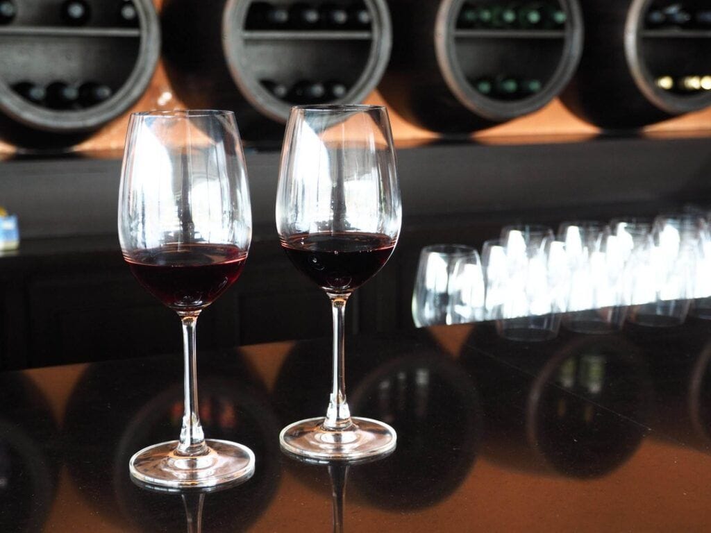 Two glasses of red wine are placed on a glossy bar counter, with shelves holding wine bottles from a sophisticated wine cellar in the background.