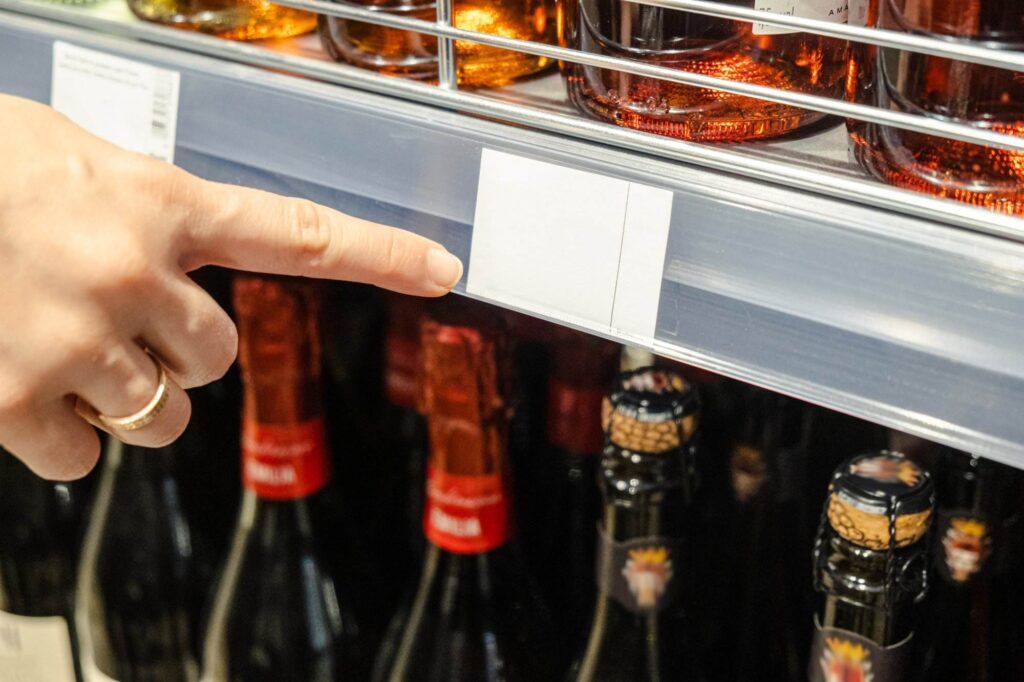 A person's hand pointing at a blank price tag on a shelf with various bottles of alcohol in a store, right beside the section for Single-Zone Wine Refrigerators.