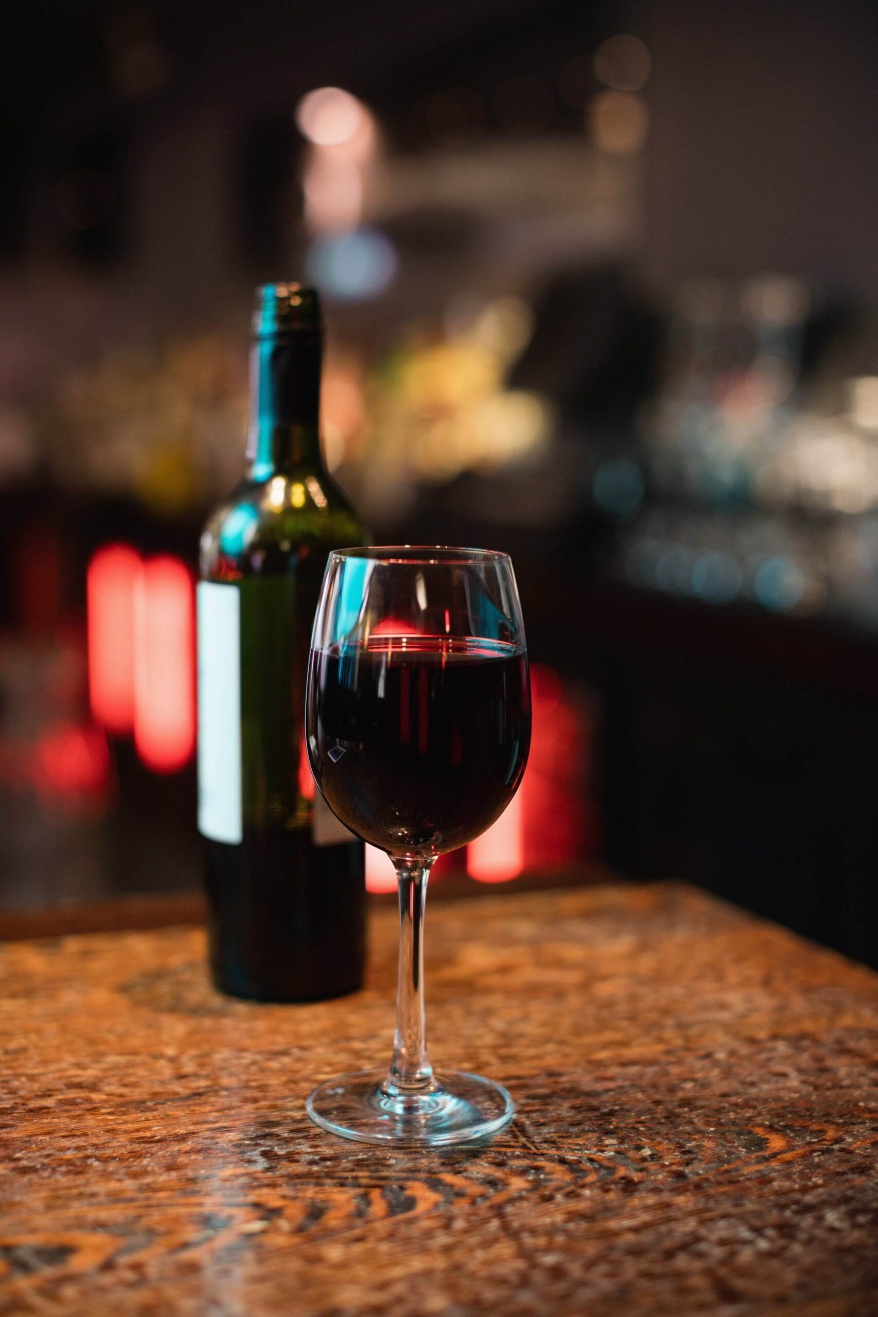 A glass of red wine sits on a wooden table with a bottle of wine in the background, set in a dimly-lit bar. Nearby, patrons discuss Wine Guardian systems, comparing models over their favorite vintages.