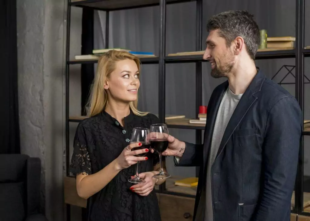 A woman and a man stand indoors holding glasses of red wine, looking at each other, with a wooden shelf and books in the background, showcasing their elegant Whisperkool wine hardware that keeps their collection perfectly chilled.