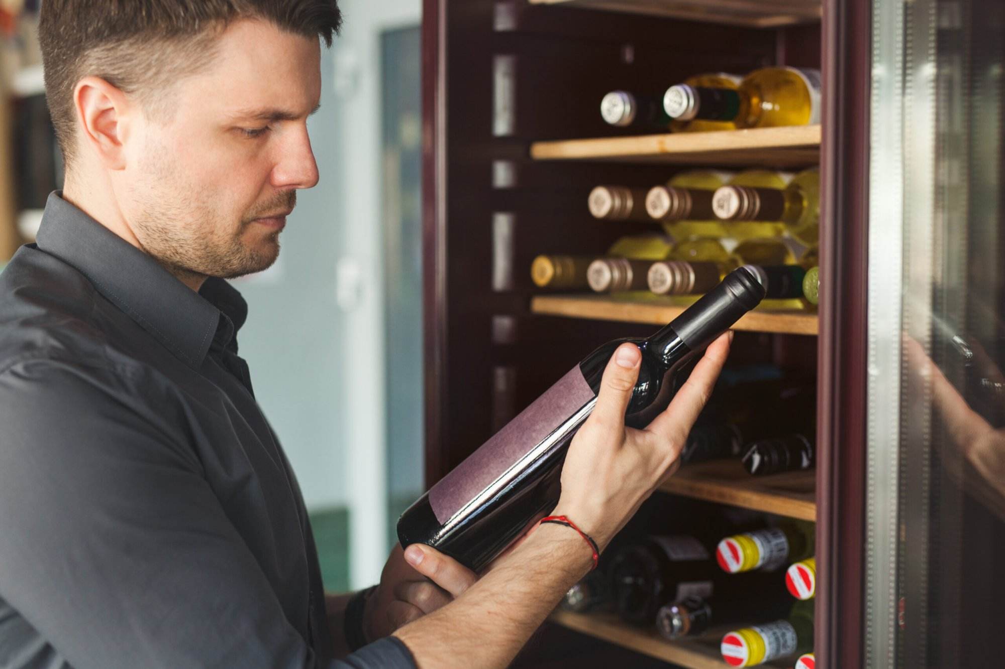 A man holding and examining a wine bottle in front of an open wine refrigerator with several bottles on the shelves, showcasing top features of modern wine hardware.