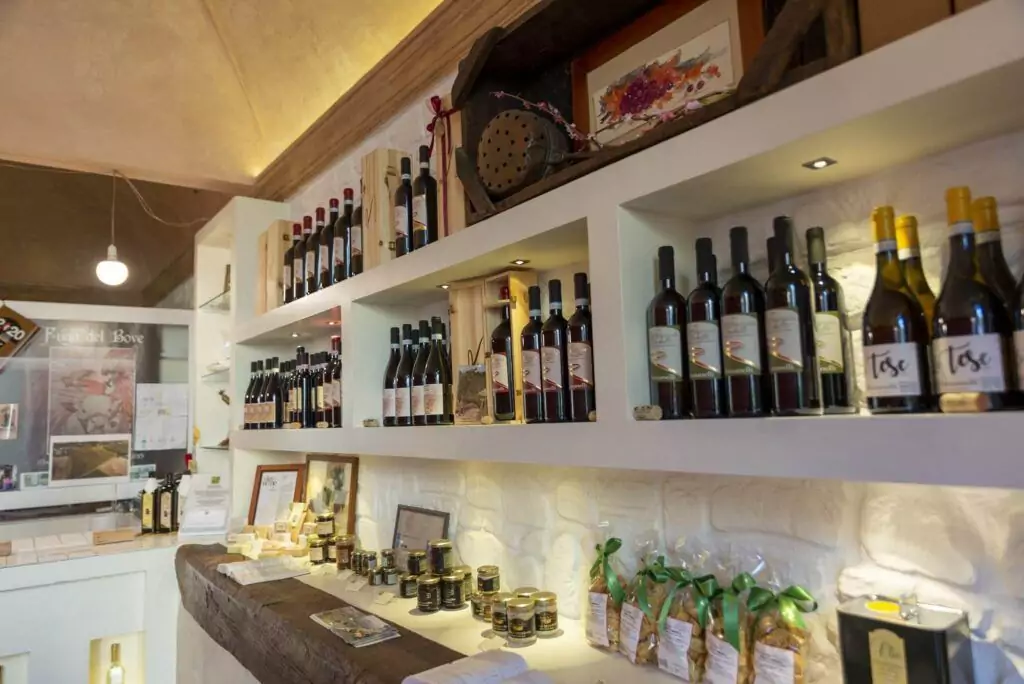 A store shelf displaying a variety of wine bottles and other gourmet food items, including jars and packaged goods. The setting is well-lit with a counter showcasing serving suggestions and additional displays in the background.