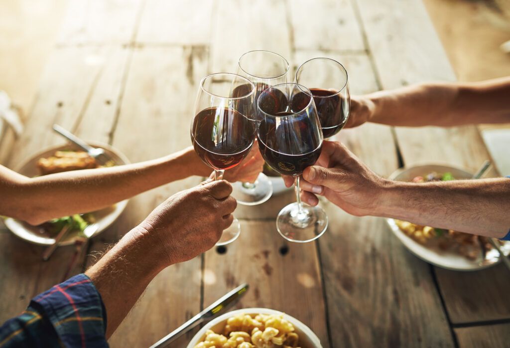 Four people clink wine glasses over a wooden table with plates of food in front of them, showcasing excellent serving suggestions and the joy of dining together.