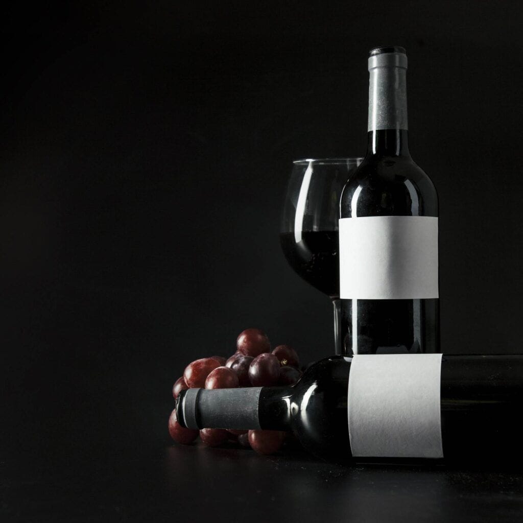 Two bottles of red wine with blank labels, one standing and one lying down, next to a full wine glass and a small bunch of red grapes, all set against a dark background—an ideal setup for showcasing your wine storage or highlighting the elegance of your Wine Guardian collection.