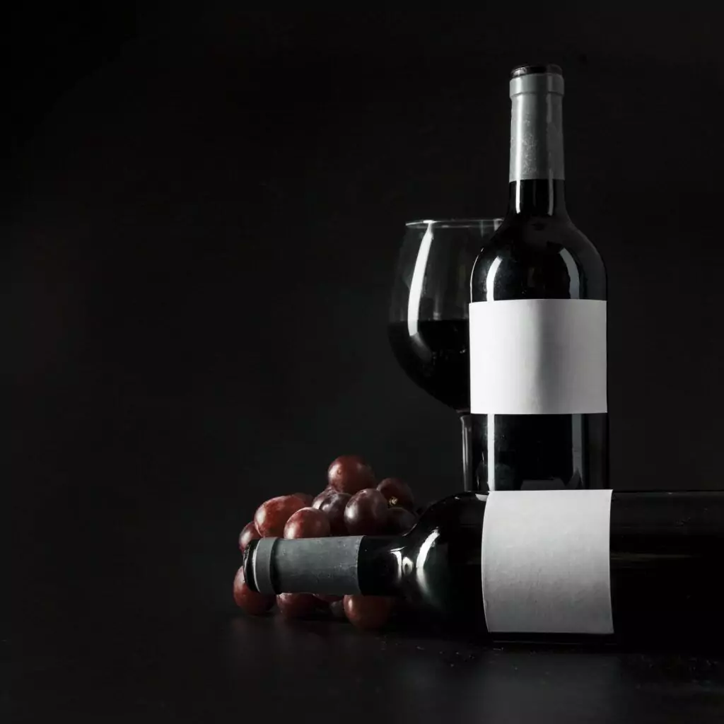 Two bottles of red wine with blank labels, one standing and one lying down, next to a full wine glass and a small bunch of red grapes, all set against a dark background—an ideal setup for showcasing your wine storage or highlighting the elegance of your Wine Guardian collection.