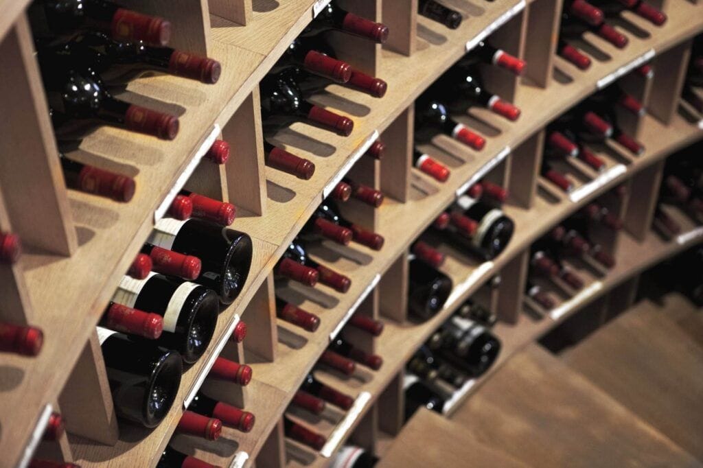 Wooden racks arranged in a curved layout, maintaining rows of wine bottles with red and black caps in a pristine wine cellar.