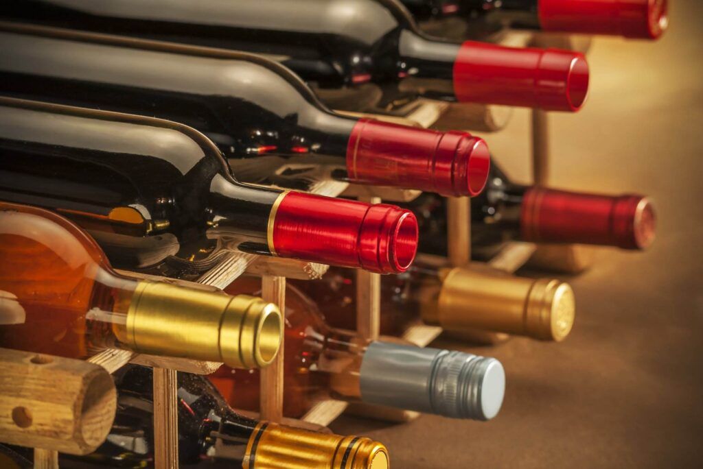 A close-up of various wine bottles with different colored caps stored horizontally in a wooden wine rack from Wine Hardware, showcasing efficient space-saving tips.