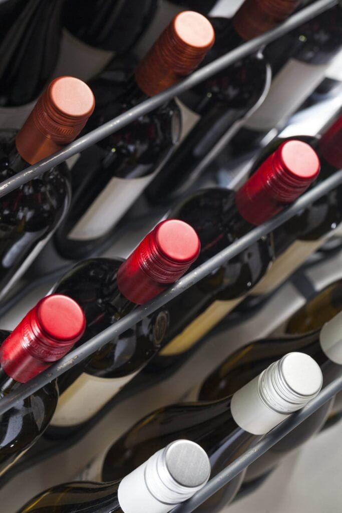 Close-up of multiple wine bottles stored on their sides in a sleek wine rack, showcasing a variety of colored caps, including red, orange, and silver. This scene perfectly captures space-saving tips and stylish wine hardware for your collection.