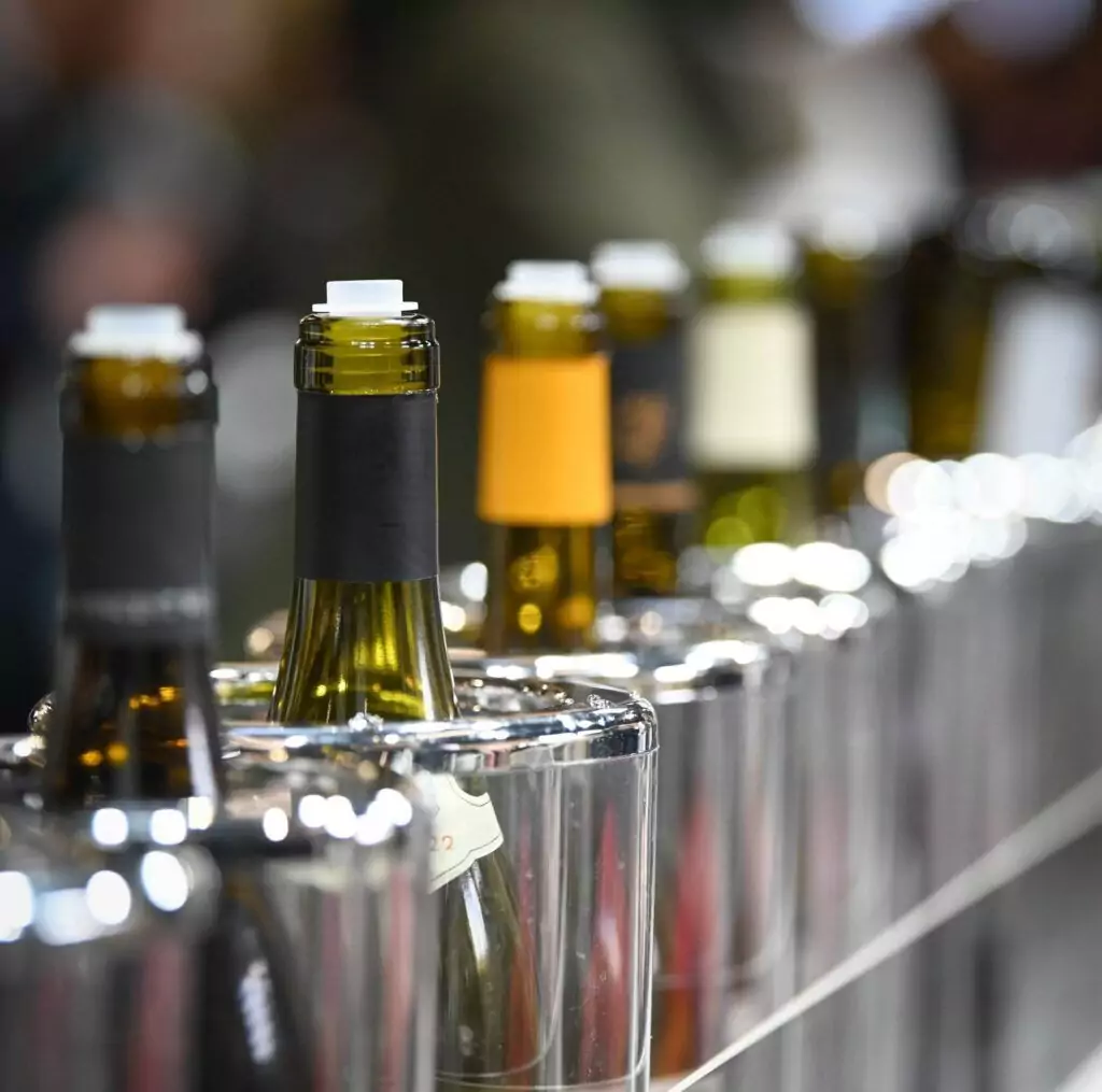 A space-saving wine rack displays a row of wine bottles with various colored labels, placed neck-down into individual stainless steel containers. The background is beautifully blurred, adding an elegant touch to any home décor.