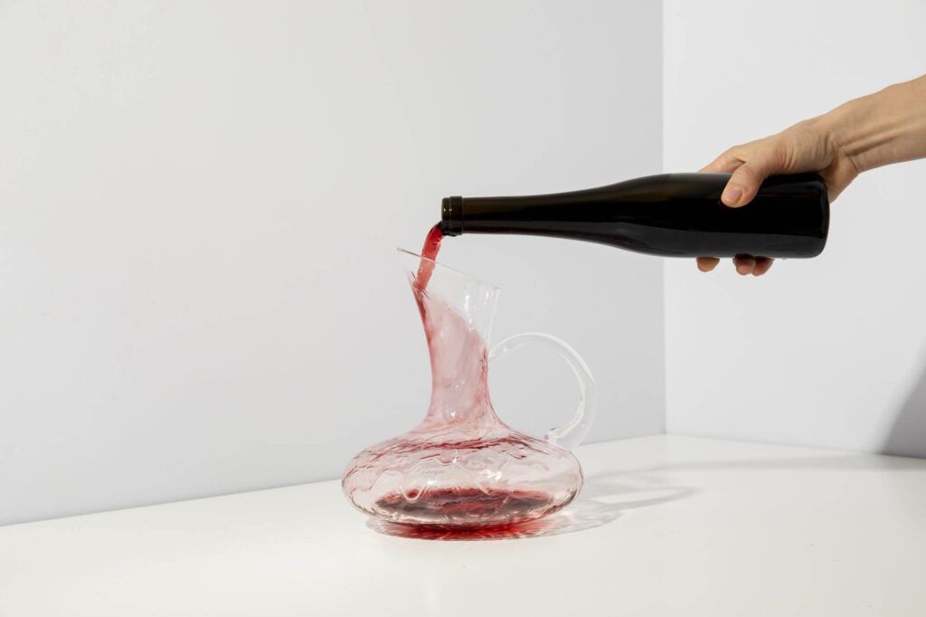 A hand is pouring red wine from a black bottle into a clear glass decanter on a white surface against a light background, highlighting the elegance of essential wine hardware.