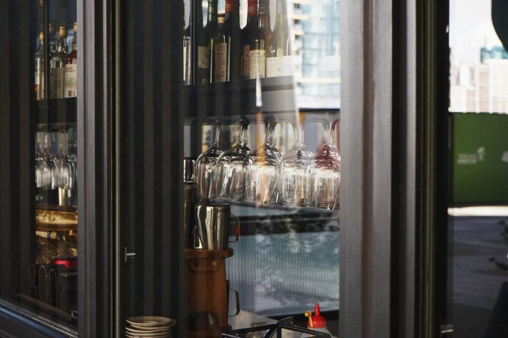 A window view reveals shelves with the best brands of wine bottles and hanging wine glasses inside a sophisticated bar, alongside sleek wine refrigerators seamlessly integrated into the design.