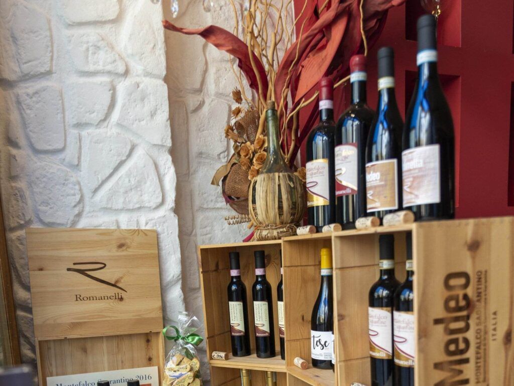 Display of wine bottles on wooden shelves, alongside wine glasses, decorative ceramic items, and imitation grapes. The backdrop features wooden boxes and a framed poster against a stone wall, complete with serving suggestions for enjoying your wine safely.