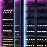 A modern wine cellar with glass doors, showcasing shelves of neatly organized bottles from the best brands, all illuminated by pink and blue LED lights.