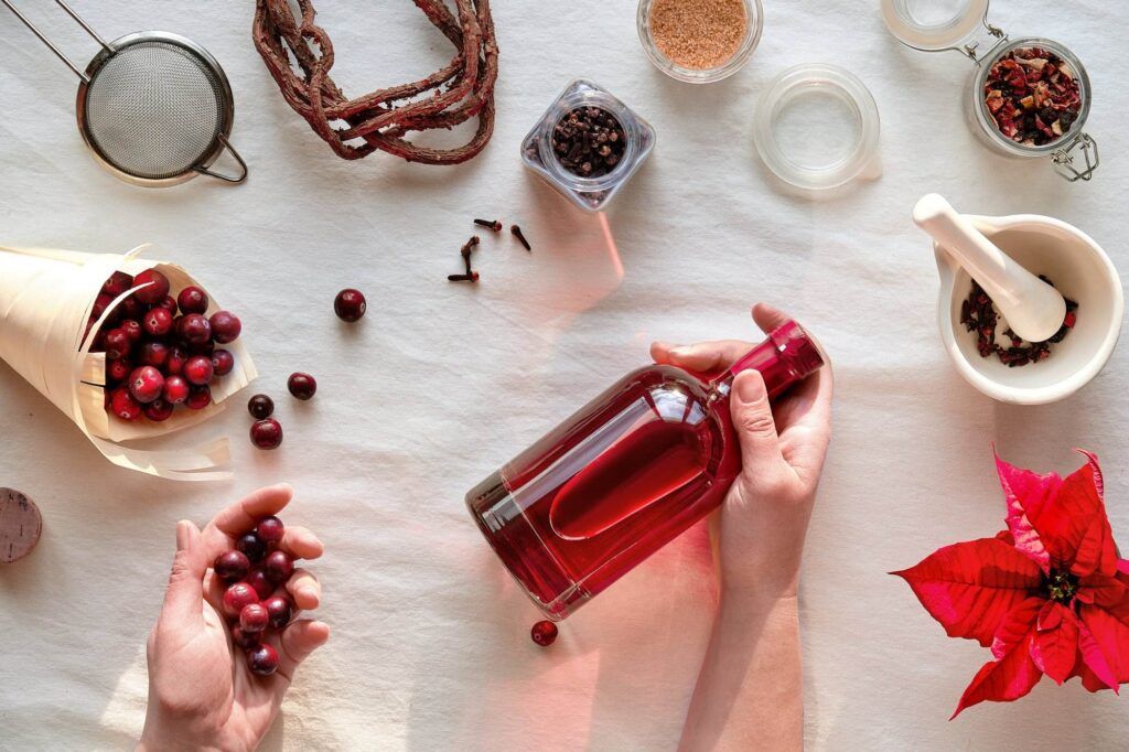 Hands holding a red bottle surrounded by cranberries, spices, a mortar and pestle, and poinsettia on a white tablecloth evoke the warmth of Walnut Creek events. Perfectly curated accessories complement the scene, reminiscent of an intimate wine tasting experience.