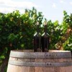 Two wine bottles on a wooden barrel in a vineyard, with lush green grapevines in the background under a clear sky.