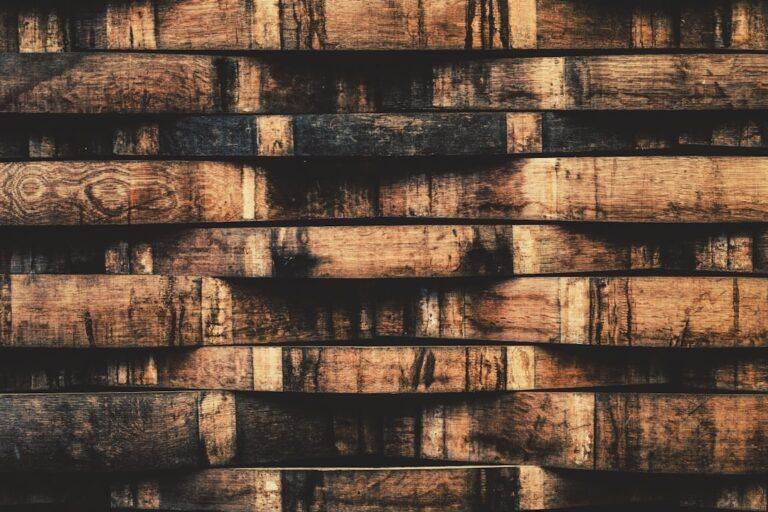 Stacked wooden barrels with a dark, aged appearance display a pattern of deep brown and black hues. The wood grain is visible, emphasizing a rustic and textured surface.