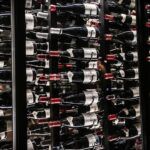 Bottles of wine stored horizontally on multiple racks in a dimly lit cellar, with a mix of red and white wine labels visible.