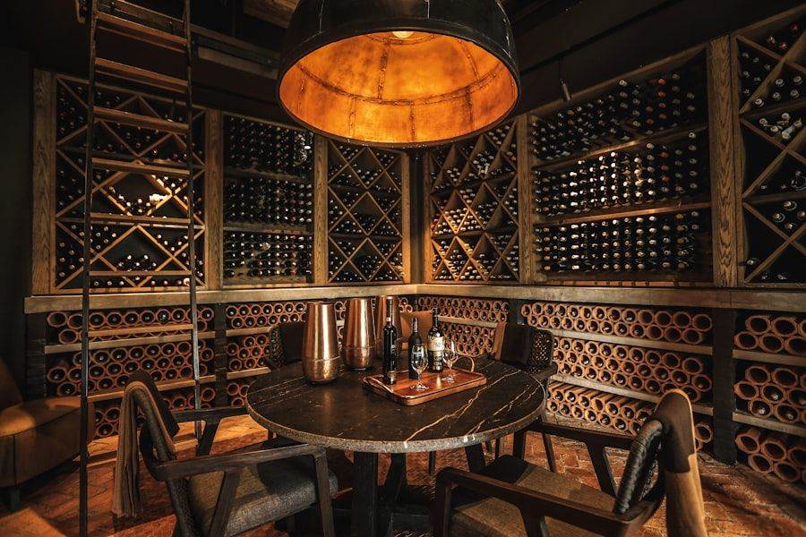 Cozy wine cellar with a round table, chairs, and bottles on top. Shelves stocked with wine bottles surround the space. Warm lighting from a large overhead lamp creates an inviting atmosphere.