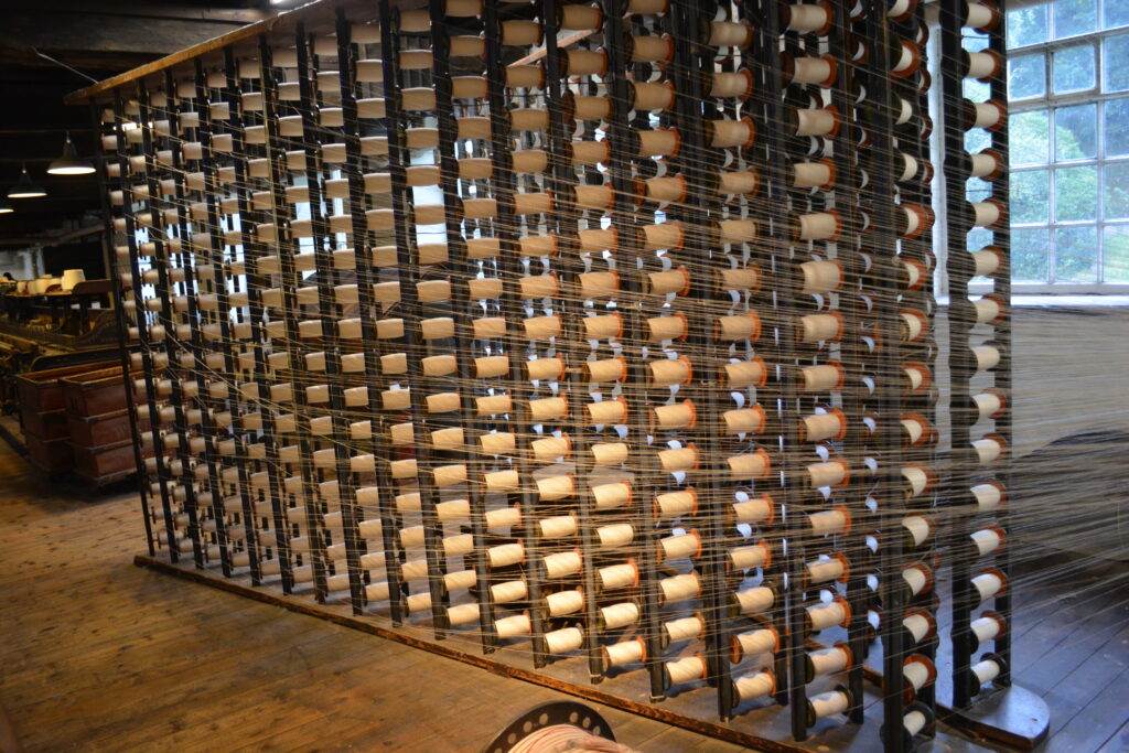 A large wooden loom with numerous spools of thread is set up in a room with wooden floors, resembling a craftsman's studio. The window on the right side offers a glimpse of Walnut Creek, blending seamlessly like architects' designs with nature.