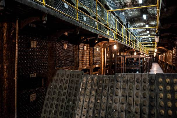 Dimly lit wine cellar in Danville with rows of wine bottles stacked on racks. Professionally designed, the space is supported by metal beams and illuminated by overhead lights, a testament to expert Wine Cellar Installers' craftsmanship.