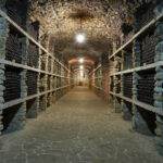 The dimly lit wine cellar in Danville features stone walls and a ceiling, elegantly showcasing rows of stacked wine bottles on wooden shelves lining the narrow aisle. Crafted by professional wine cellar installers, it exudes sophistication and charm.