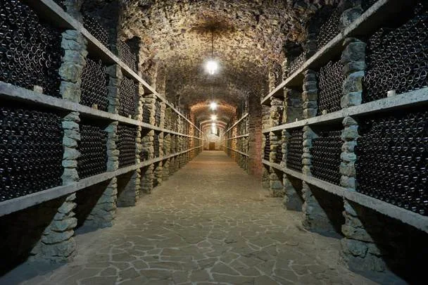 The dimly lit wine cellar in Danville features stone walls and a ceiling, elegantly showcasing rows of stacked wine bottles on wooden shelves lining the narrow aisle. Crafted by professional wine cellar installers, it exudes sophistication and charm.