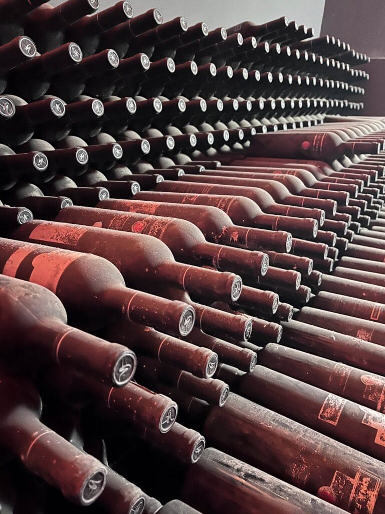 Stacks of dusty wine bottles are arranged horizontally in rows, creating a pattern in a dimly lit cellar. The scene evokes an appreciation for professional cellar construction that's built to preserve true vintages.