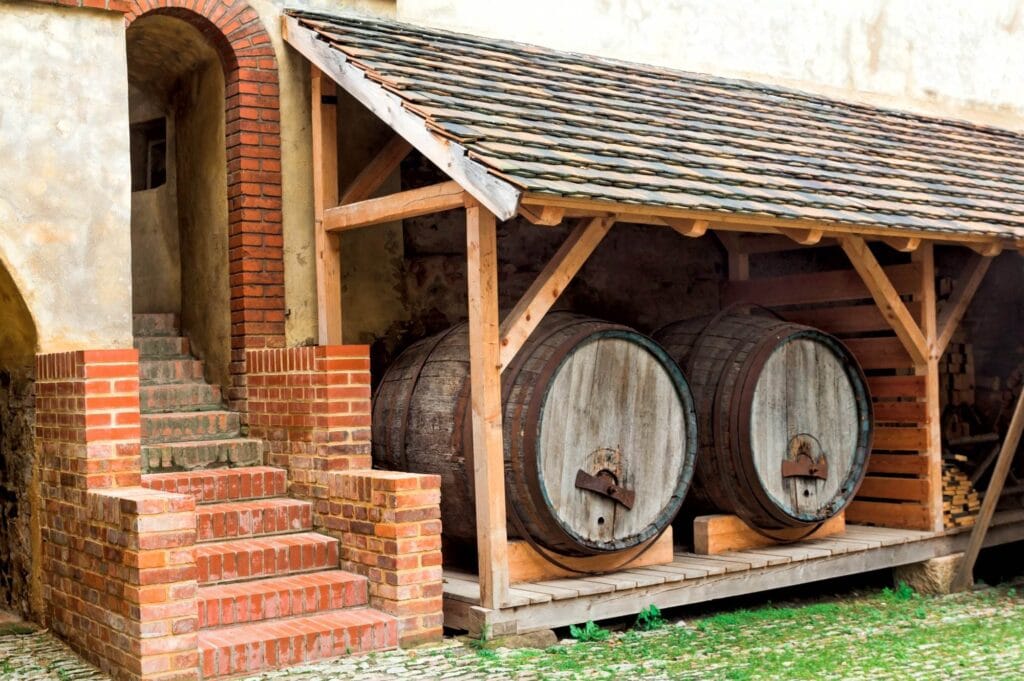 Two large wooden barrels are stored under a rustic wooden shelter next to a brick staircase against a stone building, reminiscent of outdoor wine cellars you'd find in charming Walnut Creek homes.