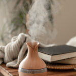 A wooden essential oil diffuser emitting vapor sits on a wooden table next to a folded blanket and two closed books, creating a cozy space where cellar humidity stays below 50 percent, with a plant in the background.