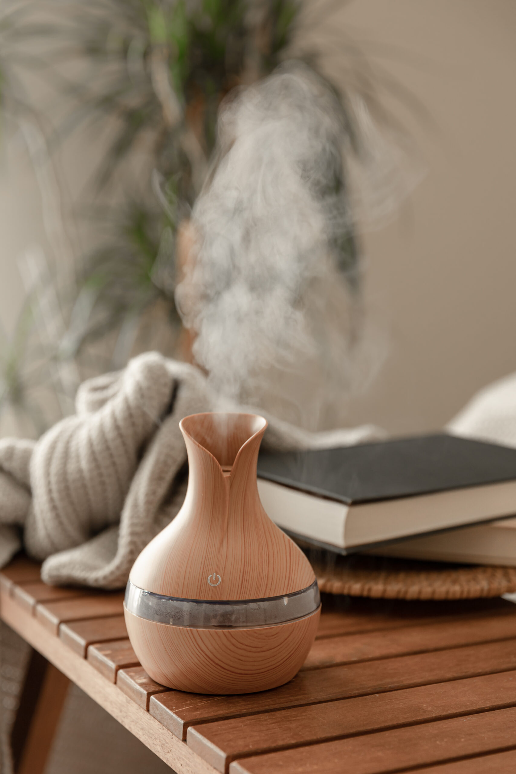 A wooden essential oil diffuser emitting vapor sits on a wooden table next to a folded blanket and two closed books, creating a cozy space where cellar humidity stays below 50 percent, with a plant in the background.