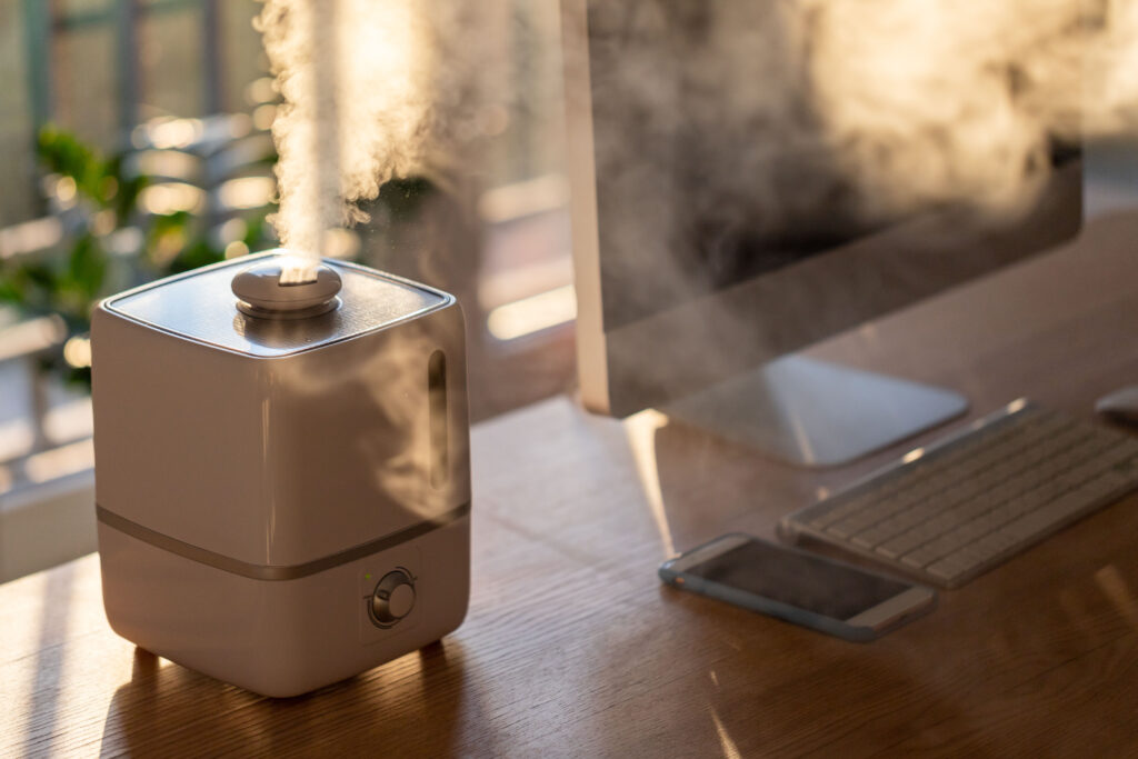 A humidifier emitting mist sits on a wooden desk next to a smartphone, keyboard, and computer monitor in natural sunlight, helping control humidity much like one would regulate cellar humidity for optimal comfort.