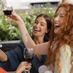 Three women smiling and raising glasses of red wine in a toast outdoors, with lush greenery and the relaxed vibe of the SF Bay Area in the background.