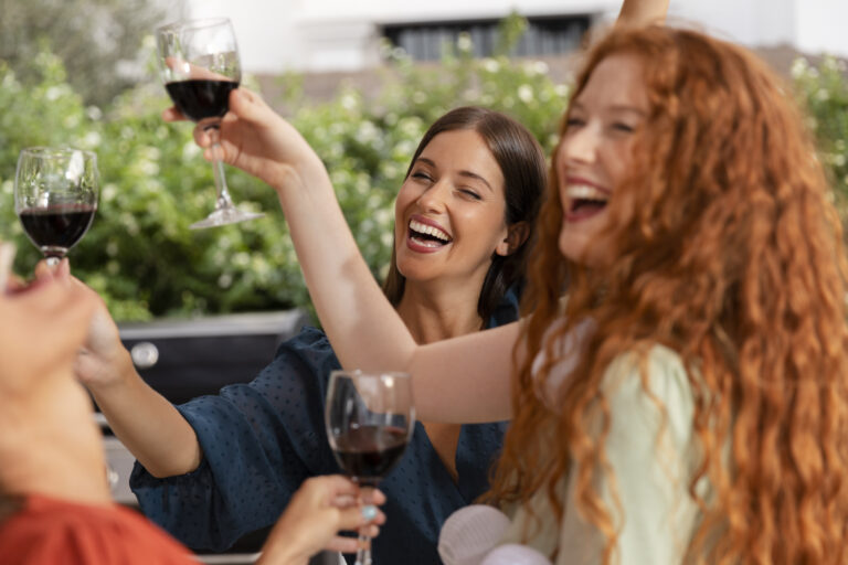 Three women smiling and raising glasses of red wine in a toast outdoors, with lush greenery and the relaxed vibe of the SF Bay Area in the background.