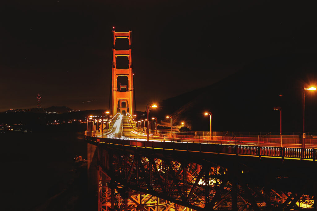 Golden Gate Bridge at night, illuminated with orange lights, spans across the dark water with vehicle light trails visible on the roadway—framed by nearby San Francisco apartments and city lights.