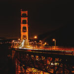 Golden Gate Bridge at night, illuminated with orange lights, spans across the dark water with vehicle light trails visible on the roadway—framed by nearby San Francisco apartments and city lights.