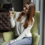 A woman with long red hair sits on a green chair by a window in the Bay Area, holding a glass of white wine and smiling as she admires it, enjoying a peaceful moment during one of the area's dry seasons.