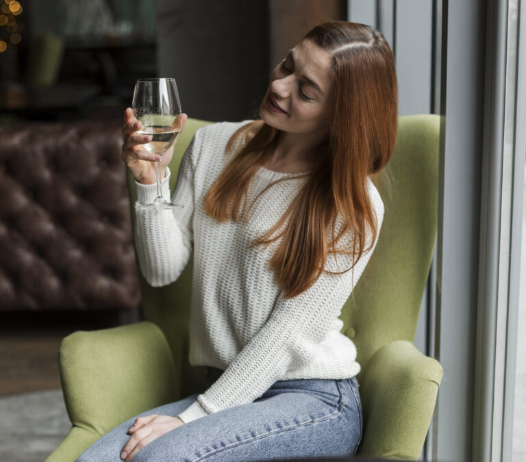 A woman with long red hair sits on a green chair by a window in the Bay Area, holding a glass of white wine and smiling as she admires it, enjoying a peaceful moment during one of the area's dry seasons.