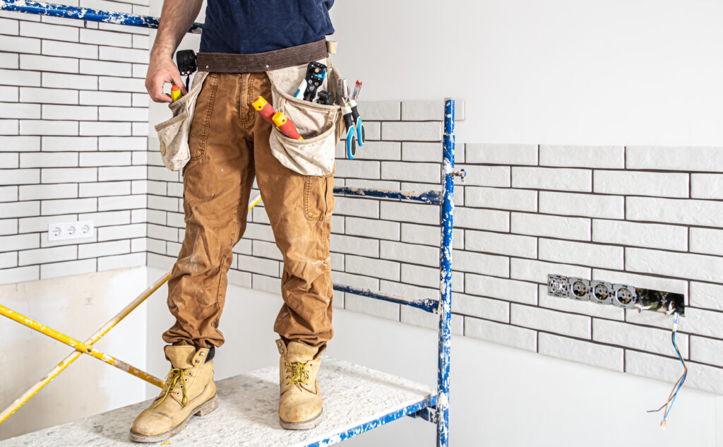 A construction worker stands on scaffolding, holding tools, with a tool belt and an unfinished brick-pattern wall—perfect for inspiring wine cellar or home remodels—in the background.