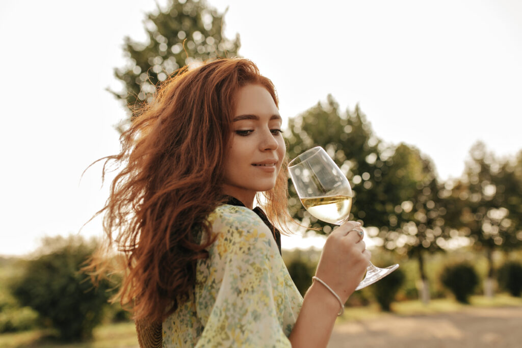 A woman with long red hair holds a glass of white wine outdoors, with green trees and sunlight in the background—perfect for those relaxed SF Bay Area afternoons or enjoying your favorite bottles from elegant wine rack kits.