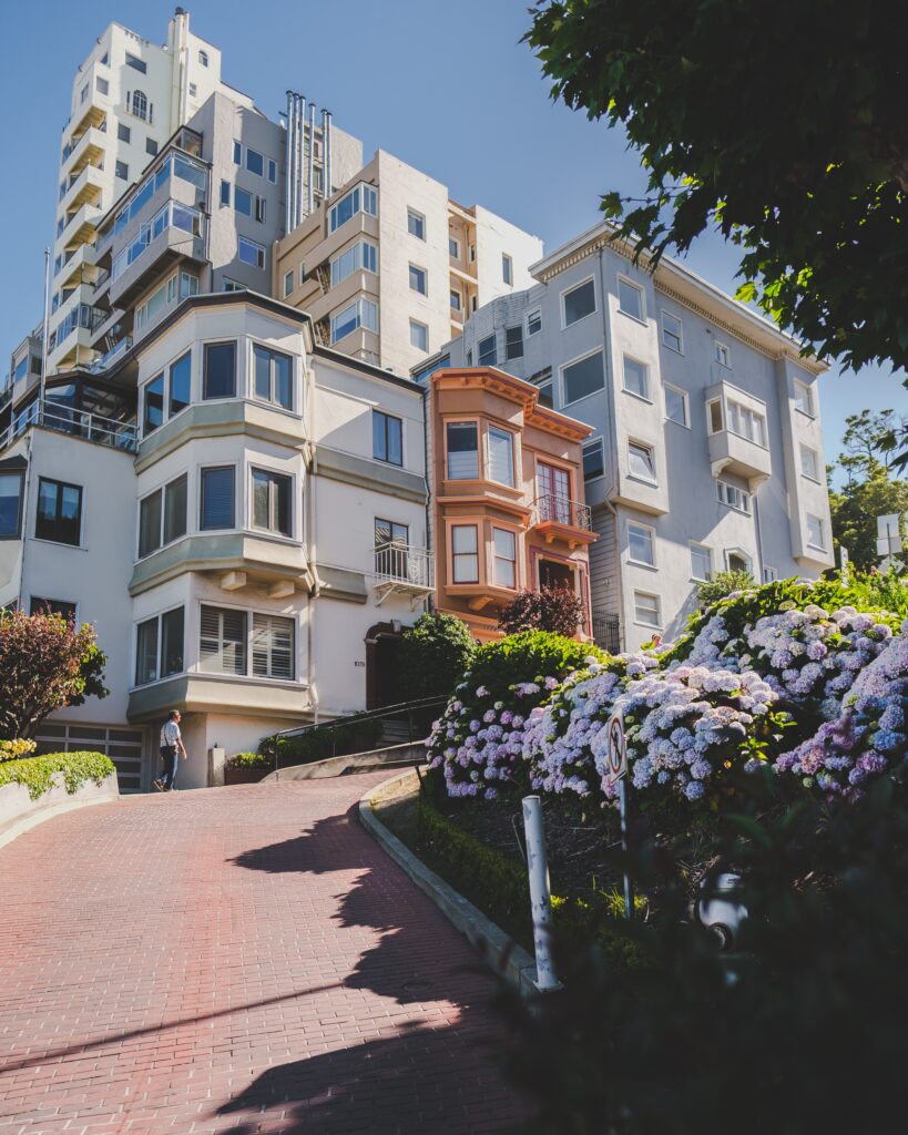 A steep, winding brick road lined with colorful hydrangeas and San Francisco apartments rises between multi-story residential buildings on a sunny day.