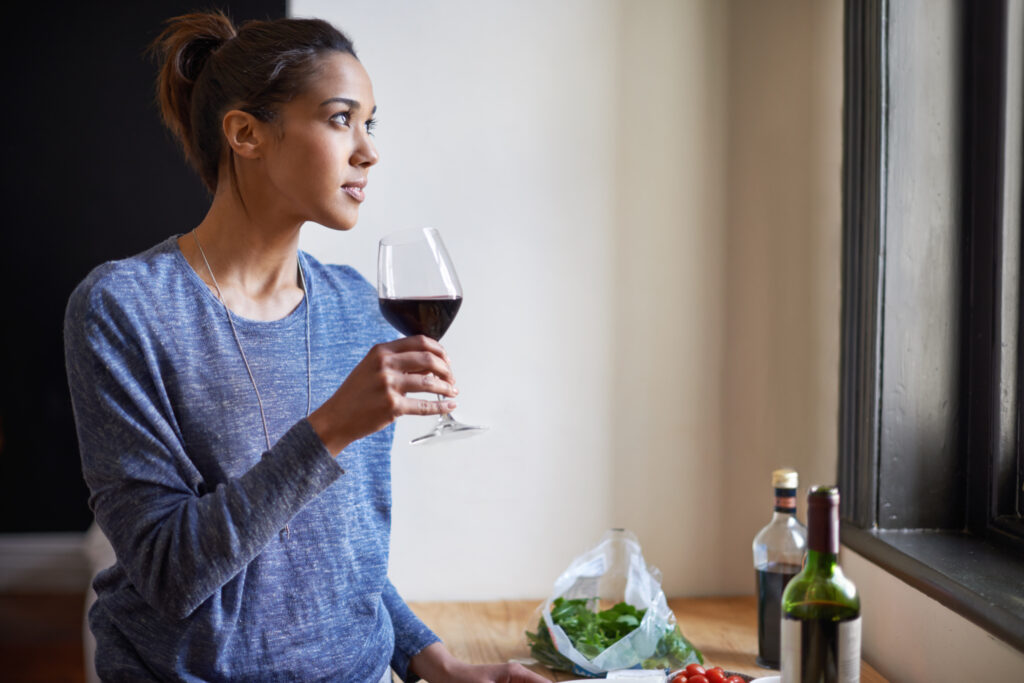 Woman in a blue shirt holds a glass of red wine, gazing out a window. On the table are salad greens, olive oil, and a wine bottle—a relaxing Bay Area scene perfect for dry seasons.