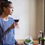 Woman in a blue shirt holds a glass of red wine, gazing out a window. On the table are salad greens, olive oil, and a wine bottle—a relaxing Bay Area scene perfect for dry seasons.