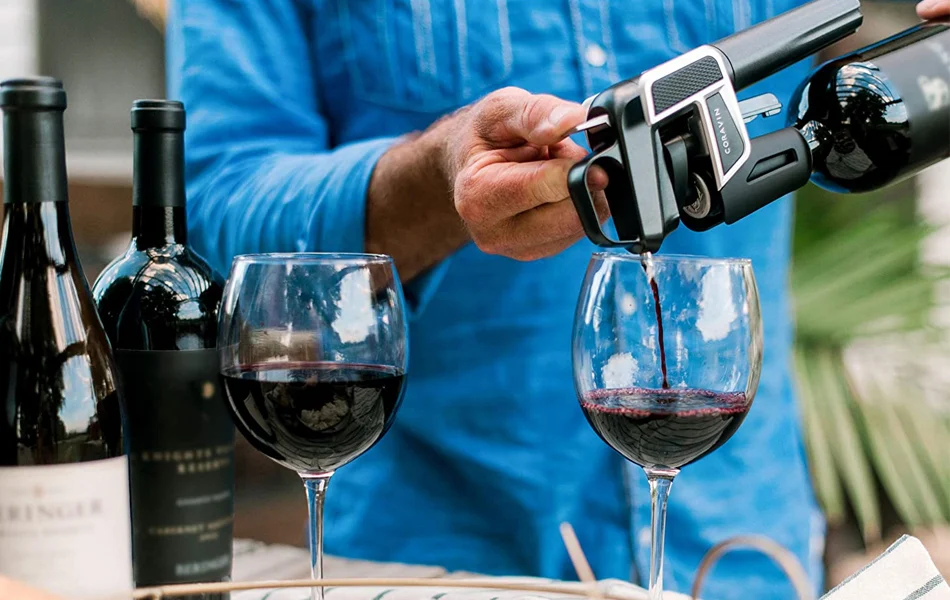 A person uses a wine preservation device with argon integration to pour red wine into a glass, while another filled glass and two wine bottles rest nearby on the table.