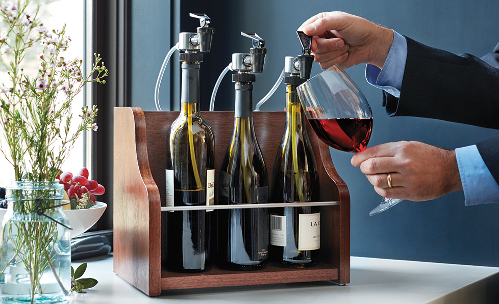 A person pours red wine from a bottle using a wine preservation system featuring argon integration, with four bottles set in a wooden rack on a countertop.